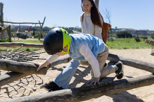 Child wearing a helmet playing on a playground climbing frame made of logs with a woman watching in the background