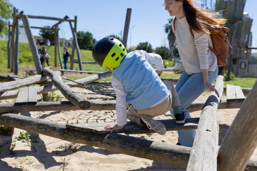Child enjoys playing on wooden playground structure while wearing a helmet, with mother supervising nearby