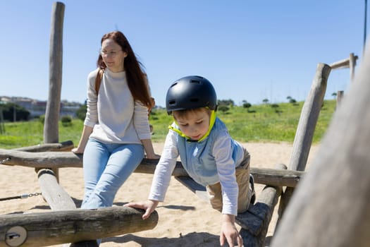 Young boy wearing protective helmet playing on playground equipment while his mother supervises