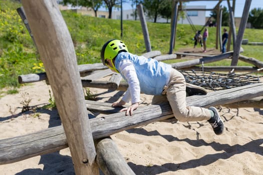 Young boy crawling on wooden beam at playground wearing protective helmet, enjoying outdoor activity