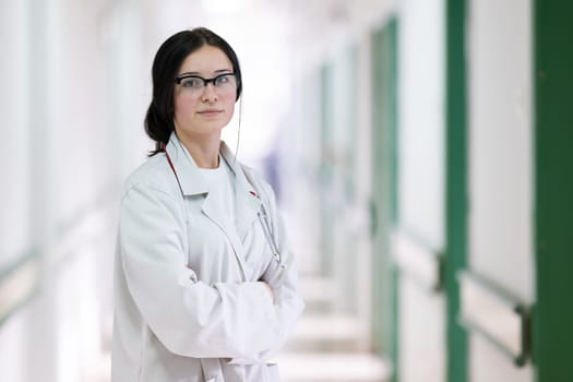Confident young female doctor with a stethoscope smiling while standing in a bright hospital corridor.