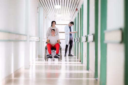 A smiling young man in a wheelchair is being pushed by a female healthcare worker in a white coat and stethoscope. They are in a hospital corridor, suggesting a medical or rehabilitation environment, evoking care and recovery.