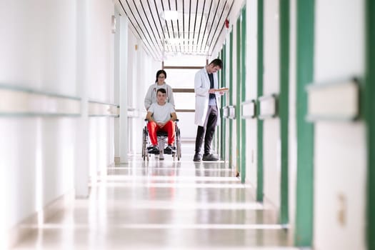 A smiling young man in a wheelchair is being pushed by a female healthcare worker in a white coat and stethoscope. They are in a hospital corridor, suggesting a medical or rehabilitation environment, evoking care and recovery.