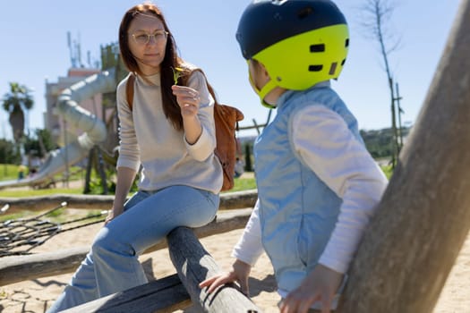 Mother showing small plant to child wearing helmet in playground