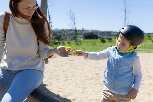 Child wearing a helmet pointing at a small yellow flower held by a woman in a park