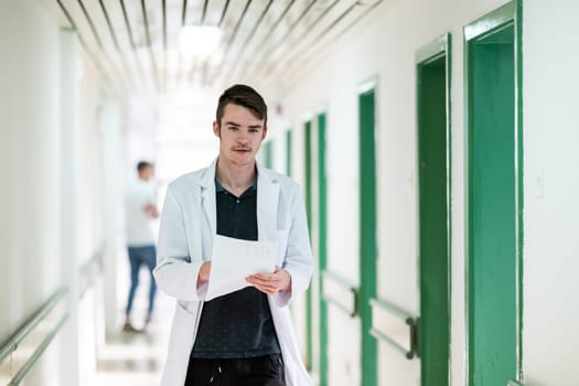 Young male doctor walking in hospital hallway while reading patient documentation. Clinical work environment.