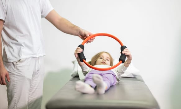 Young girl performing rehabilitation exercises with the help of a trainer, using a resistance ring while lying on a medical bench.