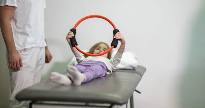 Young girl performing rehabilitation exercises with the help of a trainer, using a resistance ring while lying on a medical bench.