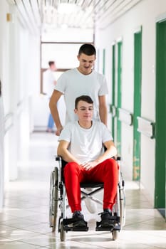 A smiling young man in a wheelchair is being pushed by a female healthcare worker in a white coat and stethoscope. They are in a hospital corridor, suggesting a medical or rehabilitation environment, evoking care and recovery.