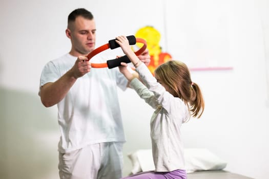 Young girl performing rehabilitation exercises with the help of a trainer, using a resistance ring while lying on a medical bench.