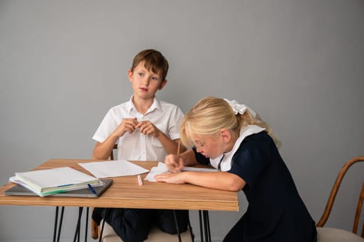A boy and a girl in school uniforms sit at a table doing homework, both focused on writing on paper.