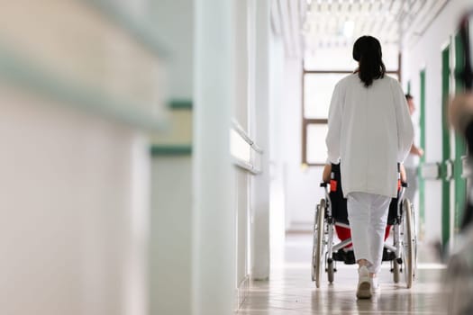 A smiling young man in a wheelchair is being pushed by a female healthcare worker in a white coat and stethoscope. They are in a hospital corridor, suggesting a medical or rehabilitation environment, evoking care and recovery.