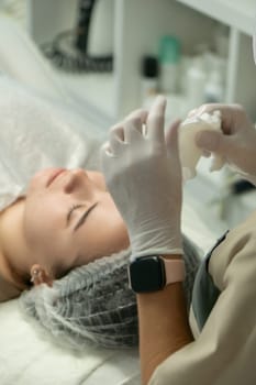 Close-up of a woman receiving a facial treatment. A gloved technician applies a cleansing or masking product to her face. Medical spa setting.