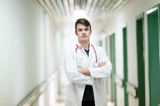 Portrait of a young doctor in white coat with arms crossed, standing in a hospital hallway.