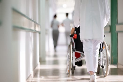 A smiling young man in a wheelchair is being pushed by a female healthcare worker in a white coat and stethoscope. They are in a hospital corridor, suggesting a medical or rehabilitation environment, evoking care and recovery.