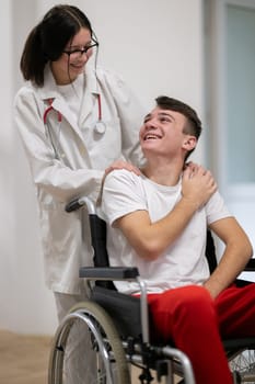 A smiling young man in a wheelchair is being pushed by a female healthcare worker in a white coat and stethoscope. They are in a hospital corridor, suggesting a medical or rehabilitation environment, evoking care and recovery.