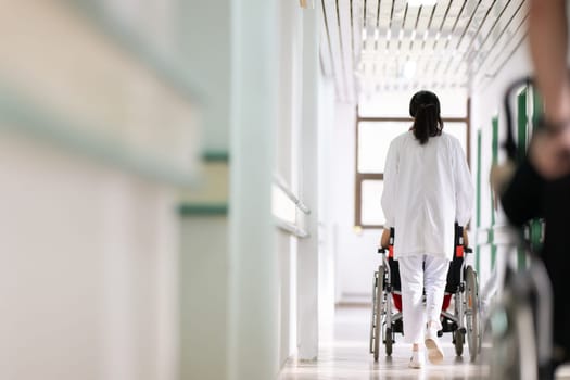 A smiling young man in a wheelchair is being pushed by a female healthcare worker in a white coat and stethoscope. They are in a hospital corridor, suggesting a medical or rehabilitation environment, evoking care and recovery.