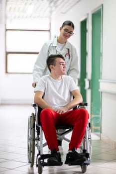 A smiling young man in a wheelchair is being pushed by a female healthcare worker in a white coat and stethoscope. They are in a hospital corridor, suggesting a medical or rehabilitation environment, evoking care and recovery.