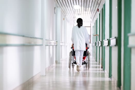 A smiling young man in a wheelchair is being pushed by a female healthcare worker in a white coat and stethoscope. They are in a hospital corridor, suggesting a medical or rehabilitation environment, evoking care and recovery.