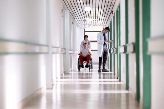 A smiling young man in a wheelchair is being pushed by a female healthcare worker in a white coat and stethoscope. They are in a hospital corridor, suggesting a medical or rehabilitation environment, evoking care and recovery.