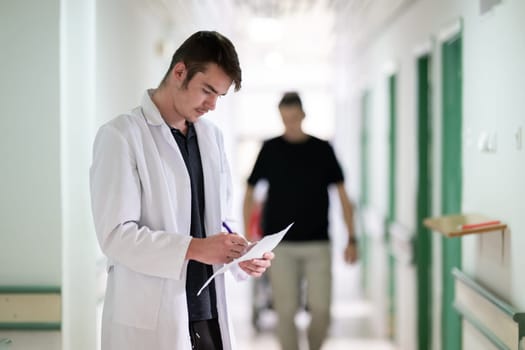Young male doctor walking in hospital hallway while reading patient documentation. Clinical work environment.