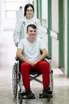 A smiling young man in a wheelchair is being pushed by a female healthcare worker in a white coat and stethoscope. They are in a hospital corridor, suggesting a medical or rehabilitation environment, evoking care and recovery.