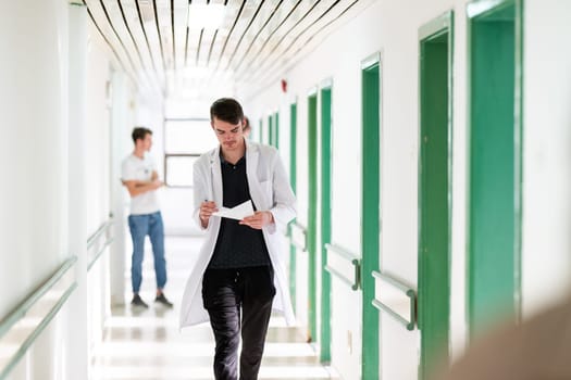 Young male doctor walking in hospital hallway while reading patient documentation. Clinical work environment.