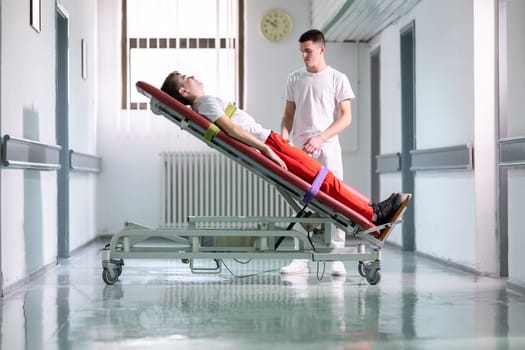Male therapist assists patient with a standing frame during a rehabilitation session in a clinical hallway. Physical recovery support concept.