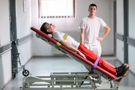 Male therapist assists patient with a standing frame during a rehabilitation session in a clinical hallway. Physical recovery support concept.