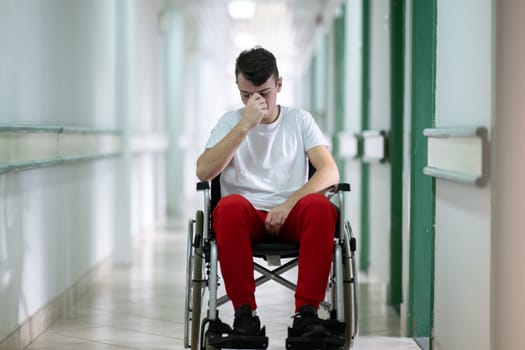 Teenage boy sitting in wheelchair with sad expression in hospital hallway. Emotion and struggle in recovery.