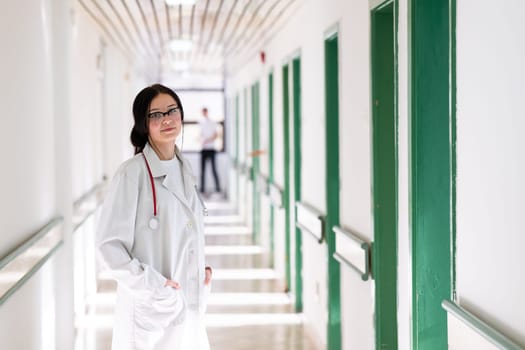 Confident young female doctor with a stethoscope smiling while standing in a bright hospital corridor.