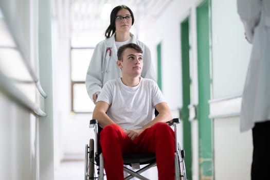 A smiling young man in a wheelchair is being pushed by a female healthcare worker in a white coat and stethoscope. They are in a hospital corridor, suggesting a medical or rehabilitation environment, evoking care and recovery.