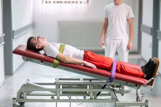 Male therapist assists patient with a standing frame during a rehabilitation session in a clinical hallway. Physical recovery support concept.