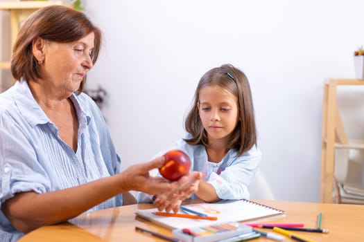 Grandmother gently places apple in girl's hand as she sits at table with colored pencils and book, creating warm, nurturing moment filled with creativity and care