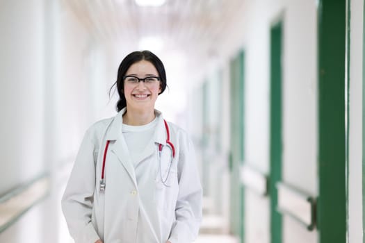 Confident young female doctor with a stethoscope smiling while standing in a bright hospital corridor.