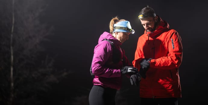 Couple night hikers examine a device under headlamp light, emphasizing navigation and teamwork under low light conditions.