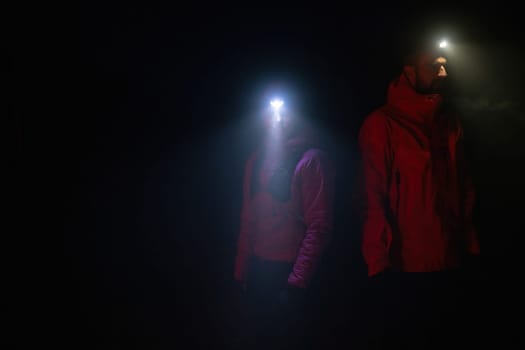 Couple equipped with headlamps explore a forest in total darkness, with beams of light cutting through the night air.