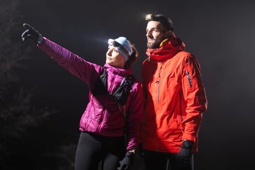 A woman points into the distance while discussing directions with her hiking partner under the beam of headlamps