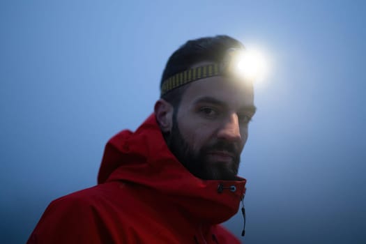 A lone hiker balances on a rocky mountain trail during early morning or dusk, with a headlamp lighting the misty path.
