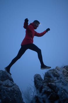 A lone hiker balances on a rocky mountain trail during early morning or dusk, with a headlamp lighting the misty path.