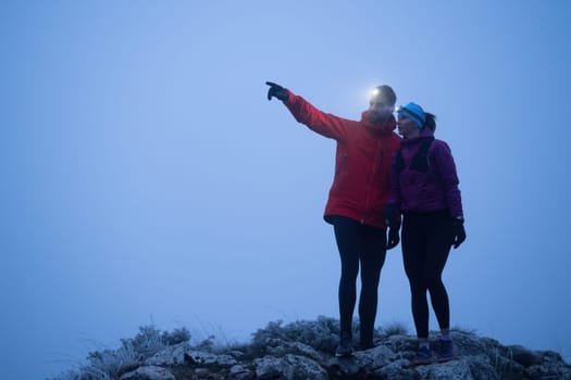Two hikers consult their GPS device at the summit of a mountain during early morning with headlamps on, surrounded by mist.