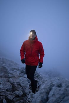 A lone hiker balances on a rocky mountain trail during early morning or dusk, with a headlamp lighting the misty path.