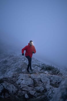 A lone hiker balances on a rocky mountain trail during early morning or dusk, with a headlamp lighting the misty path.