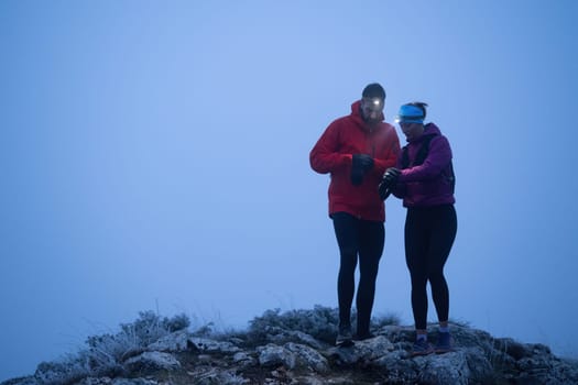Two hikers consult their GPS device at the summit of a mountain during early morning with headlamps on, surrounded by mist.