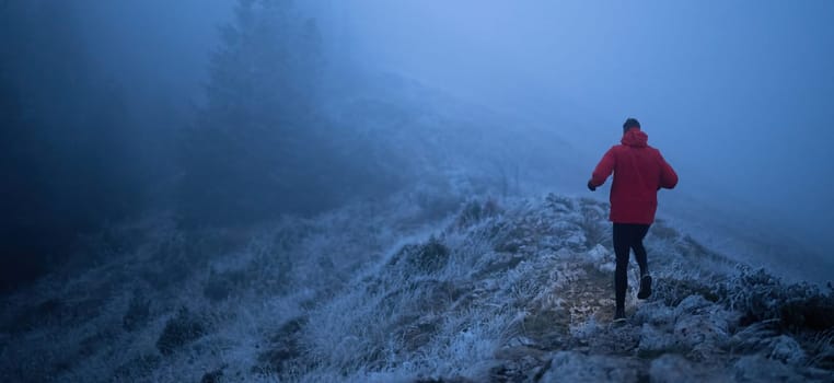 A lone hiker balances on a rocky mountain trail during early morning or dusk, with a headlamp lighting the misty path.