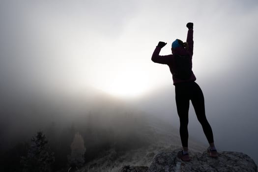 Woman celebrating on rocky mountaintop with arms raised in victory surrounded by fog.