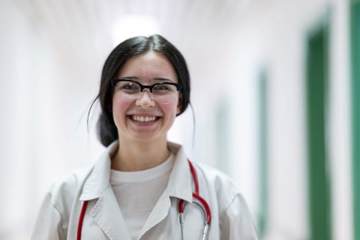 Confident young female doctor with a stethoscope smiling while standing in a bright hospital corridor.