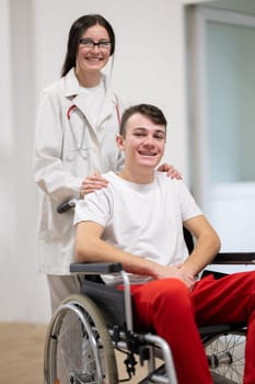 A smiling young man in a wheelchair is being pushed by a female healthcare worker in a white coat and stethoscope. They are in a hospital corridor, suggesting a medical or rehabilitation environment, evoking care and recovery.