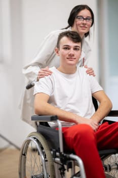 A smiling young man in a wheelchair is being pushed by a female healthcare worker in a white coat and stethoscope. They are in a hospital corridor, suggesting a medical or rehabilitation environment, evoking care and recovery.