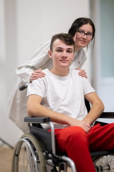 A smiling young man in a wheelchair is being pushed by a female healthcare worker in a white coat and stethoscope. They are in a hospital corridor, suggesting a medical or rehabilitation environment, evoking care and recovery.